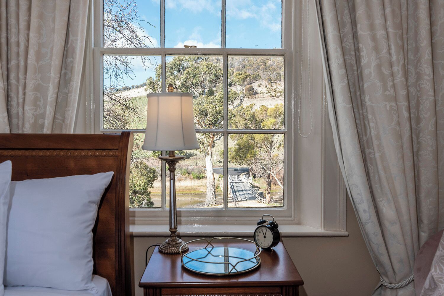 Ein Schlafzimmer mit hölzernem Kopfteil, einem Nachttisch mit Lampe, einer Uhr und Blick auf eine ländliche Landschaft durch ein Fenster.
