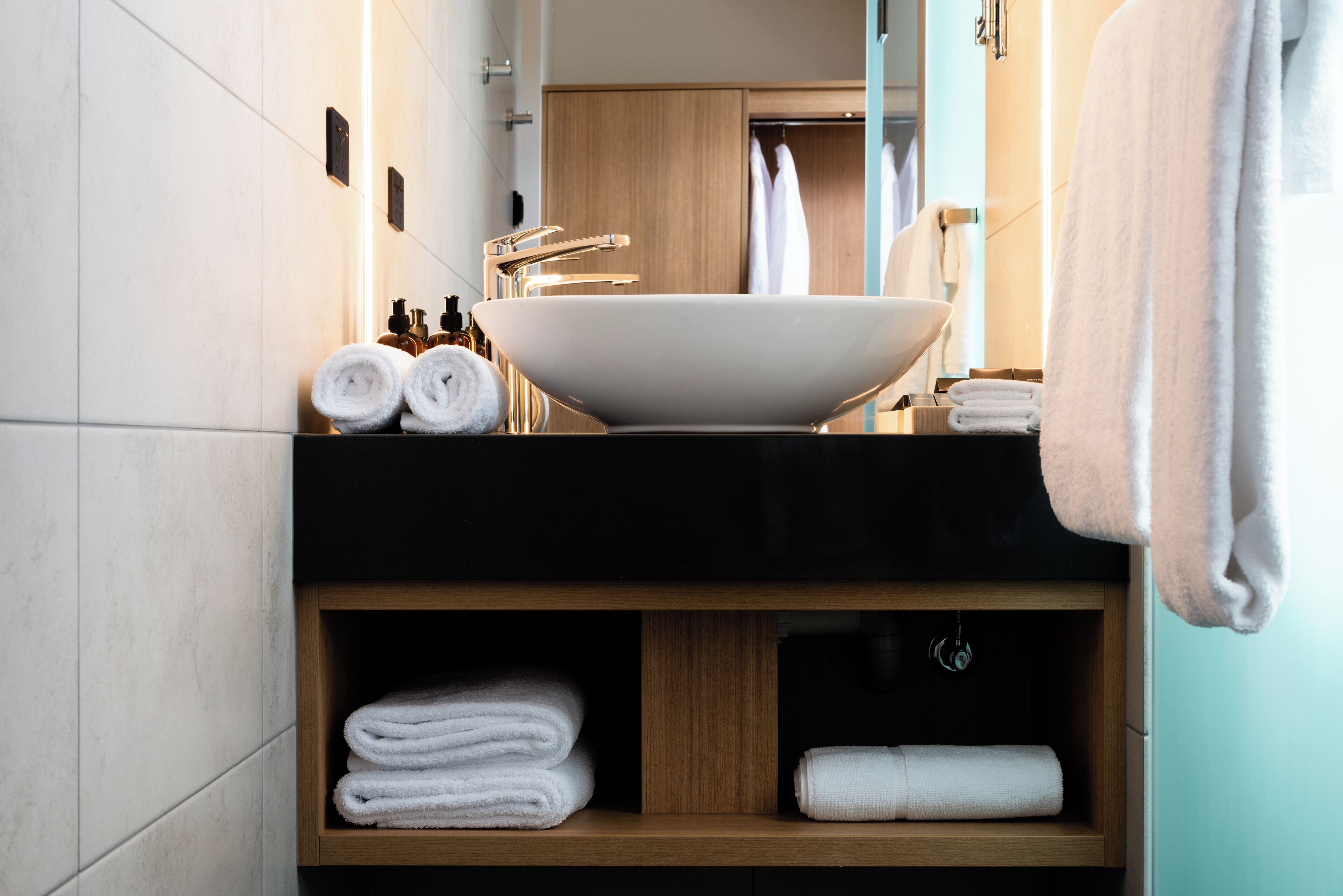 Un baño moderno con un lavamanos grande de color blanco, estantes de madera con toallas dobladas y un espejo que refleja un armario y toallas.