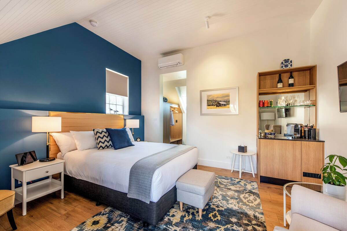 A modern hotel room with a large bed, bedside tables, a small seating area, and a kitchenette.
