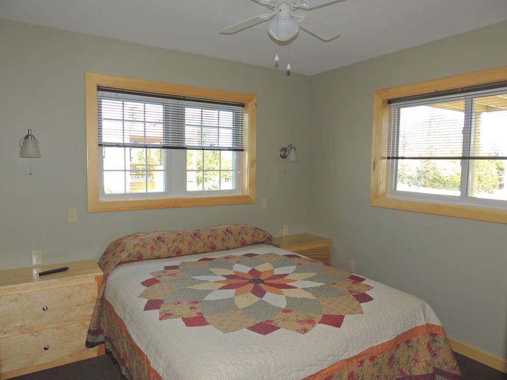 A bedroom with a bed, two windows with blinds, a ceiling fan, and a nightstand.