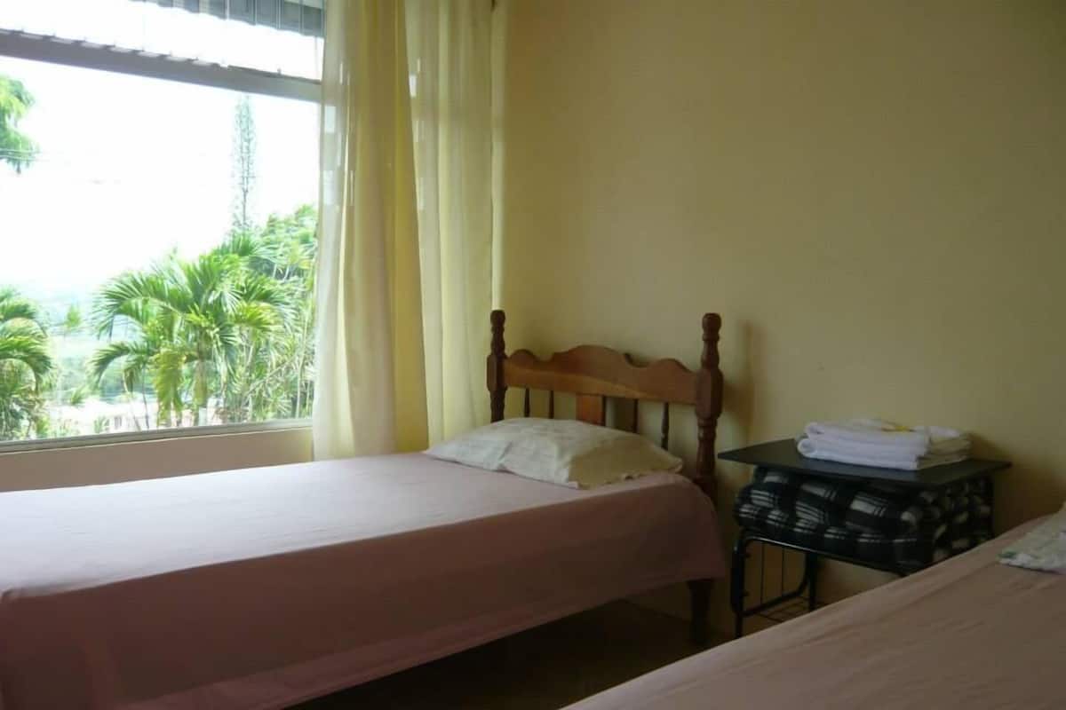 Ein Zimmer mit zwei Einzelbetten, einem kleinen Tisch mit Handtüchern und einem Fenster mit Vorhängen.