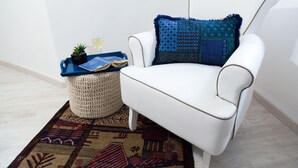A white armchair with a patterned cushion, a small side table with a tray, a potted plant, and a woven ottoman.