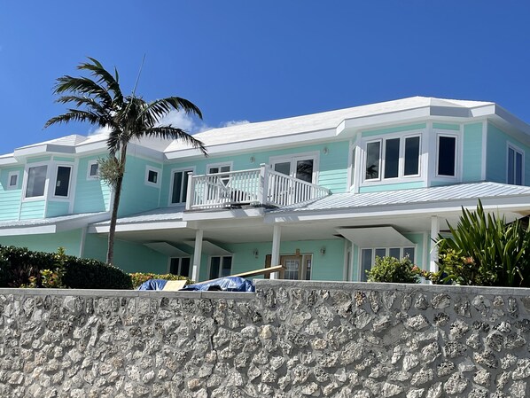 A two-story house with a balcony, a stone wall in the foreground, and a clear blue sky.