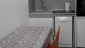 Et lille køkken med hvide skabe, en vask, komfur og opvaskemaskine. Der er et bord med dug og to stole.