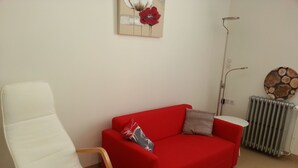 Et værelse med en hvid stol, en rød sofa, et lille hvidt bord, en radiator og et billedværk på væggen.