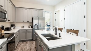 A modern kitchen with grey cabinets, a stainless steel refrigerator, a double sink, and a wooden dining table.