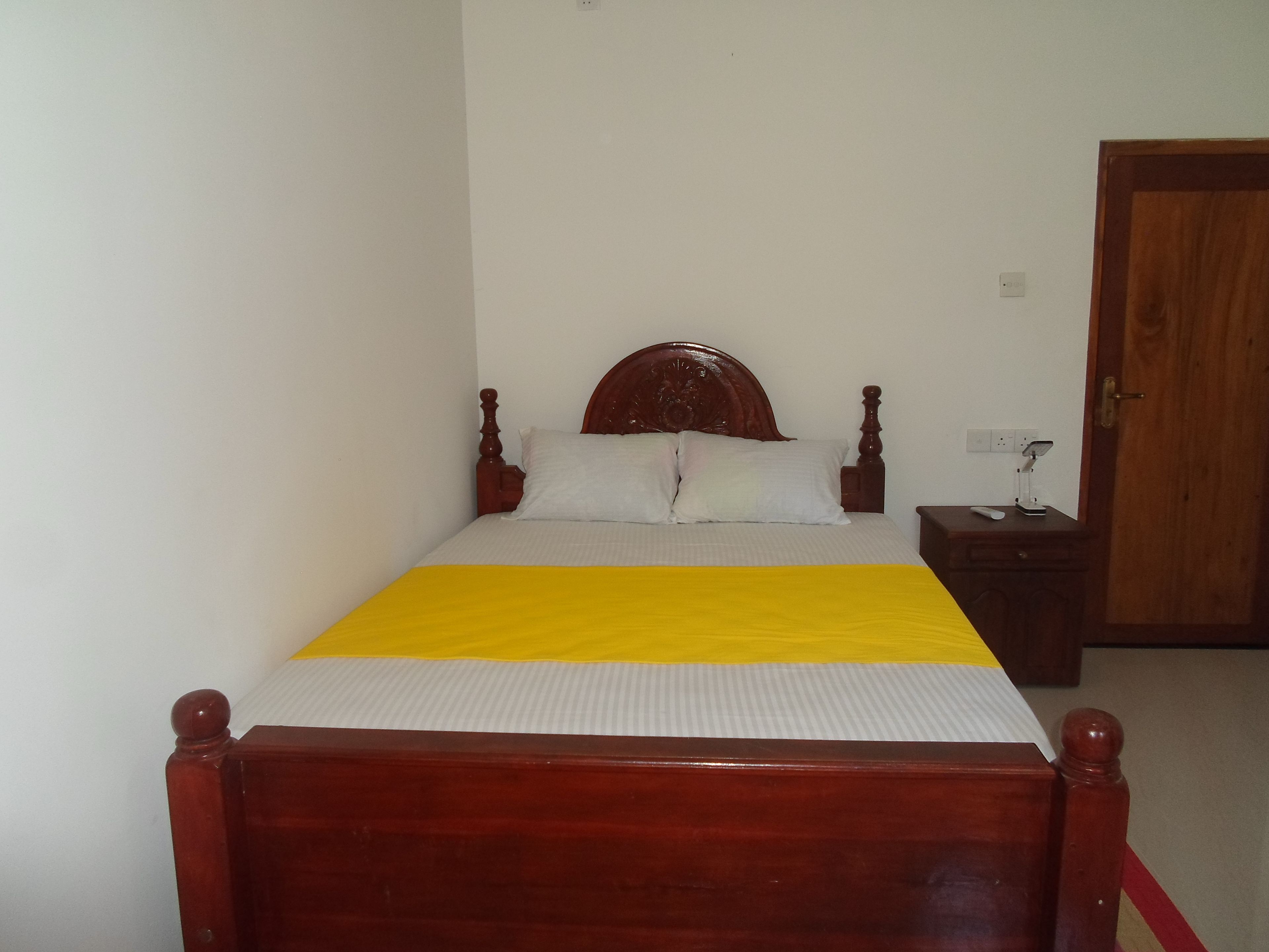 Een bed met een houten hoofdbord, wit beddengoed en een gele accentkleur. Er staat een nachtkastje met een lamp en een afstandsbediening.