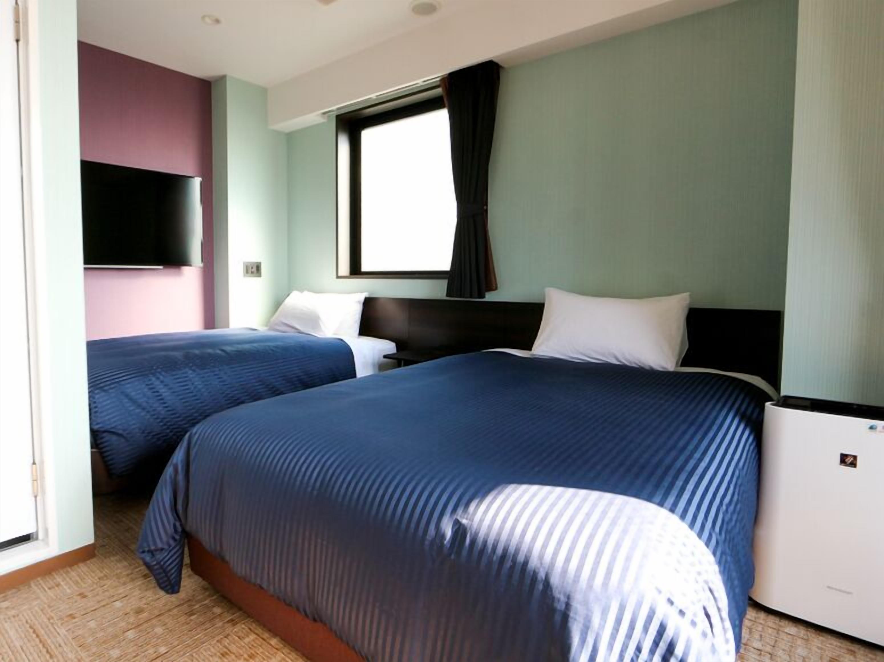 A hotel room with two beds, a flat-screen TV, a window with curtains, and a built-in air conditioning unit.