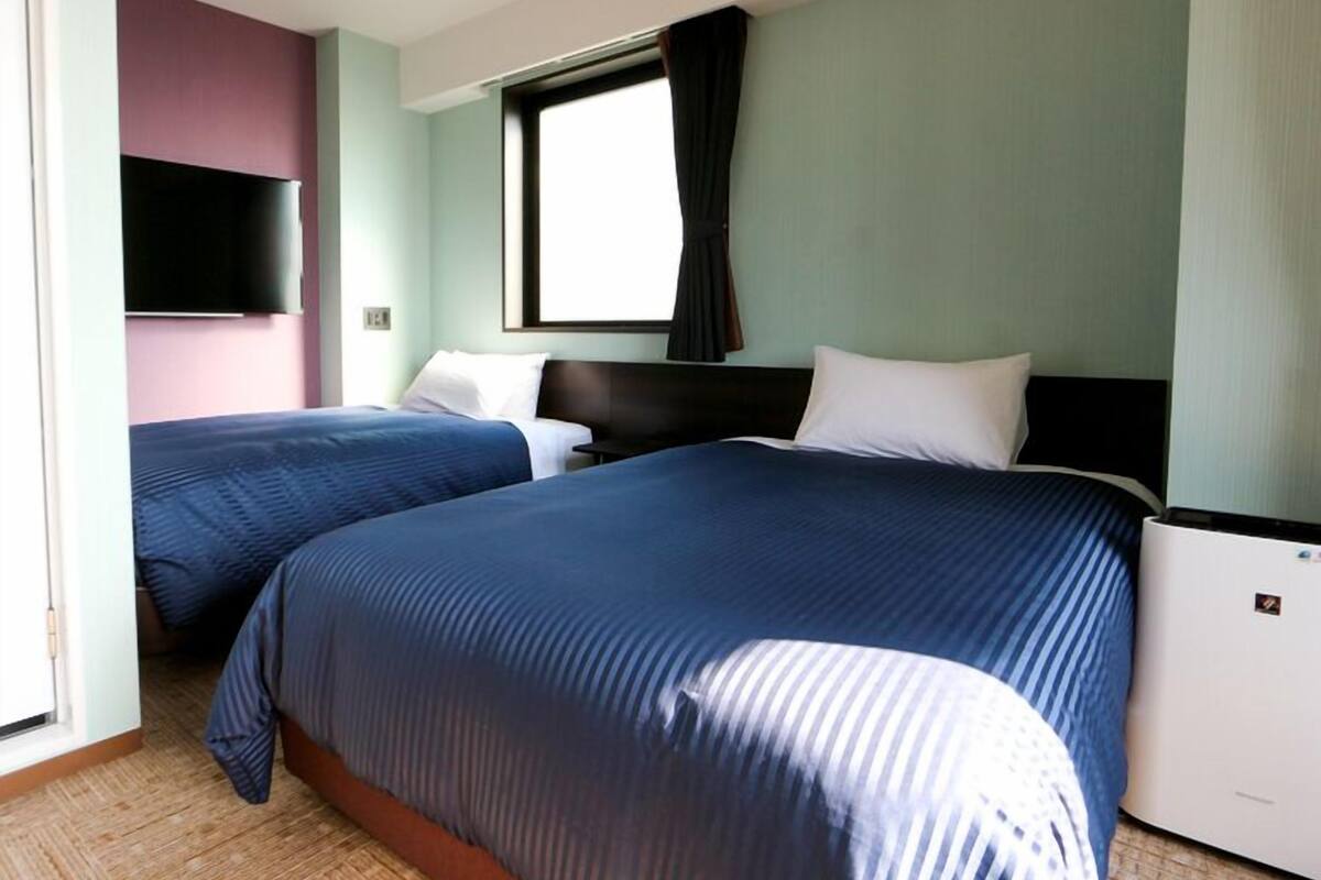 A hotel room with two beds, a flat-screen TV, a window with curtains, and a built-in air conditioning unit.