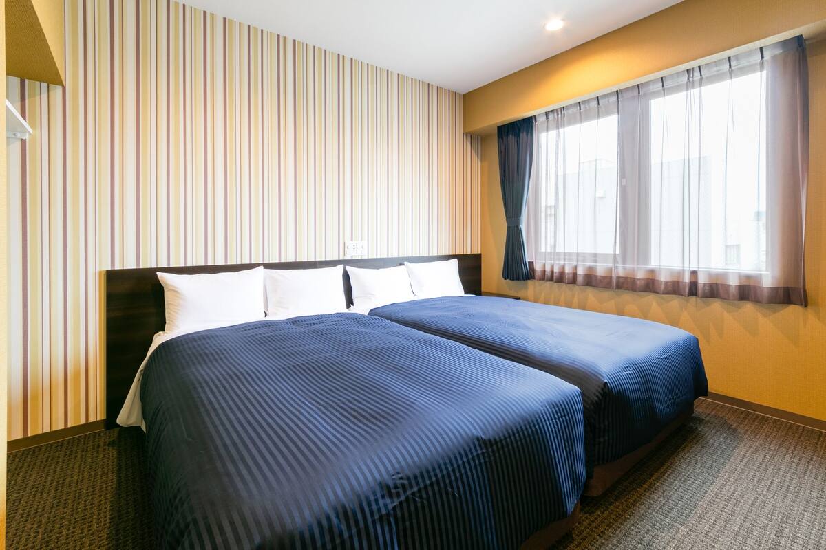 A hotel room with two beds, striped wallpaper, a window with curtains, and a carpeted floor.