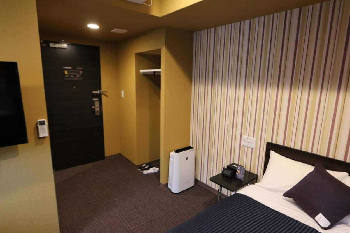 A hotel room with a bed, a nightstand, a trash bin, a coat rack, and a door.