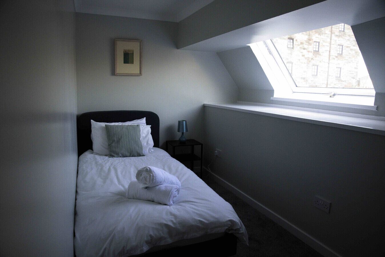 Une chambre à coucher comprenant un lit, une table de chevet, une lampe, un tableau encadré au mur et une fenêtre de toit.
