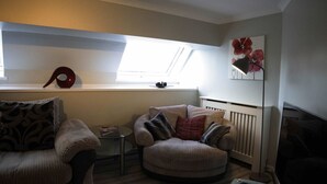A cozy living room with a sofa, armchair, and a small table. There's a radiator, a window with a view, and a painting on the wall.