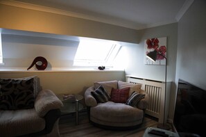 A cozy living room with a sofa, armchair, and a small table. There's a radiator, a window with a view, and a painting on the wall. - The Stables: Fabulous stylish flat in Merchant City & Parking (Glasgow)