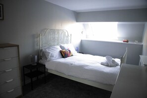 Un lit bien fait, avec du linge de lit blanc, une petite table de chevet avec une lampe, une commode et une fenêtre donnant sur l’extérieur.