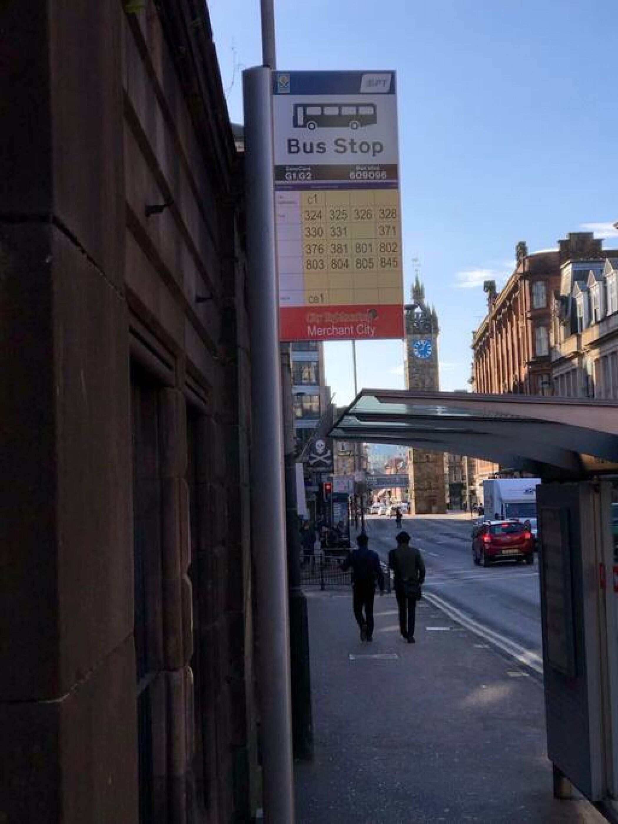 A bus stop with a schedule board, a clock tower, and a pedestrian walkway.