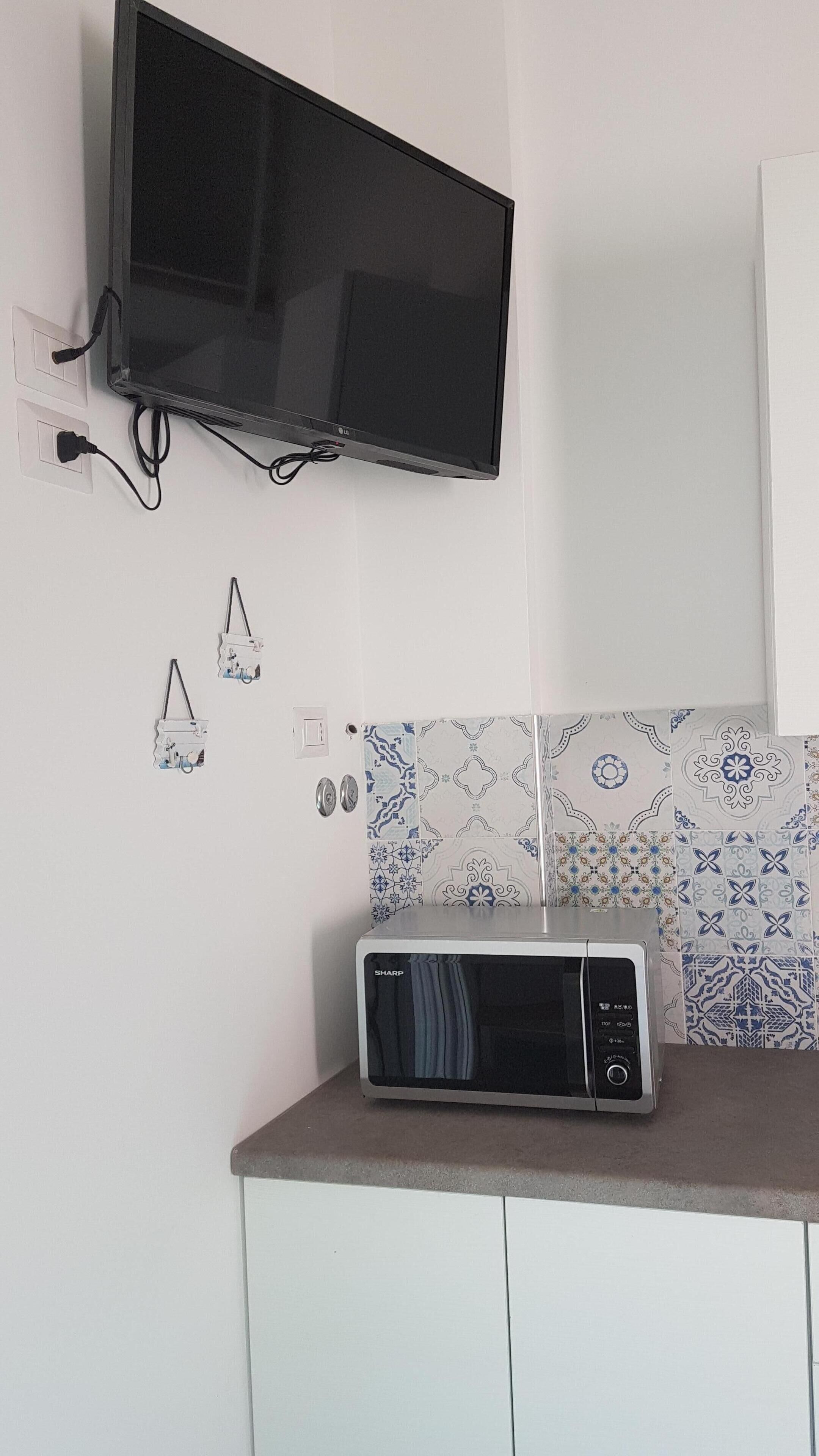 Una cocina con un televisor de pantalla plana en la pared, un microondas en la encimera y azulejos decorativos en el respuesto.