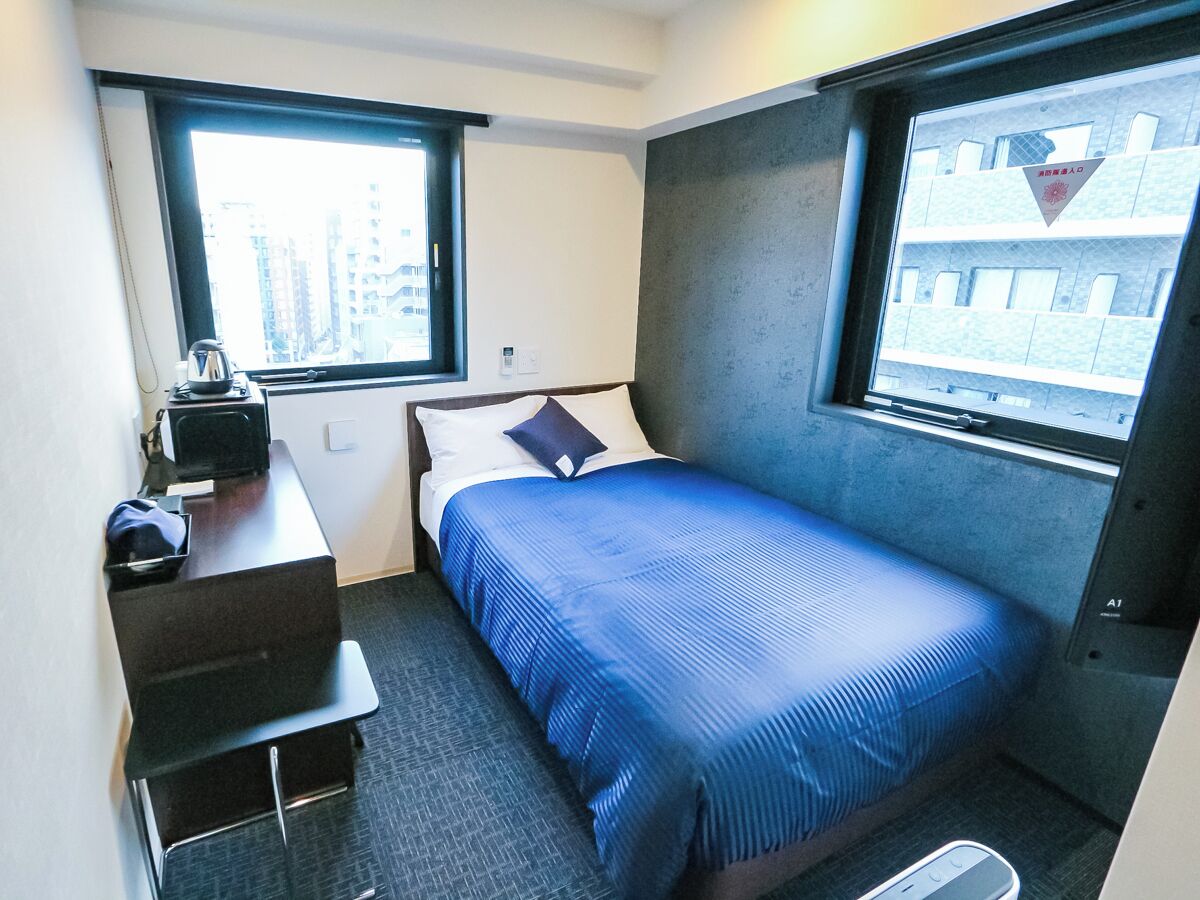 Habitación de hotel pequeña con una cama individual, un escritorio y una silla. Dos ventanas ofrecen vistas a la ciudad.