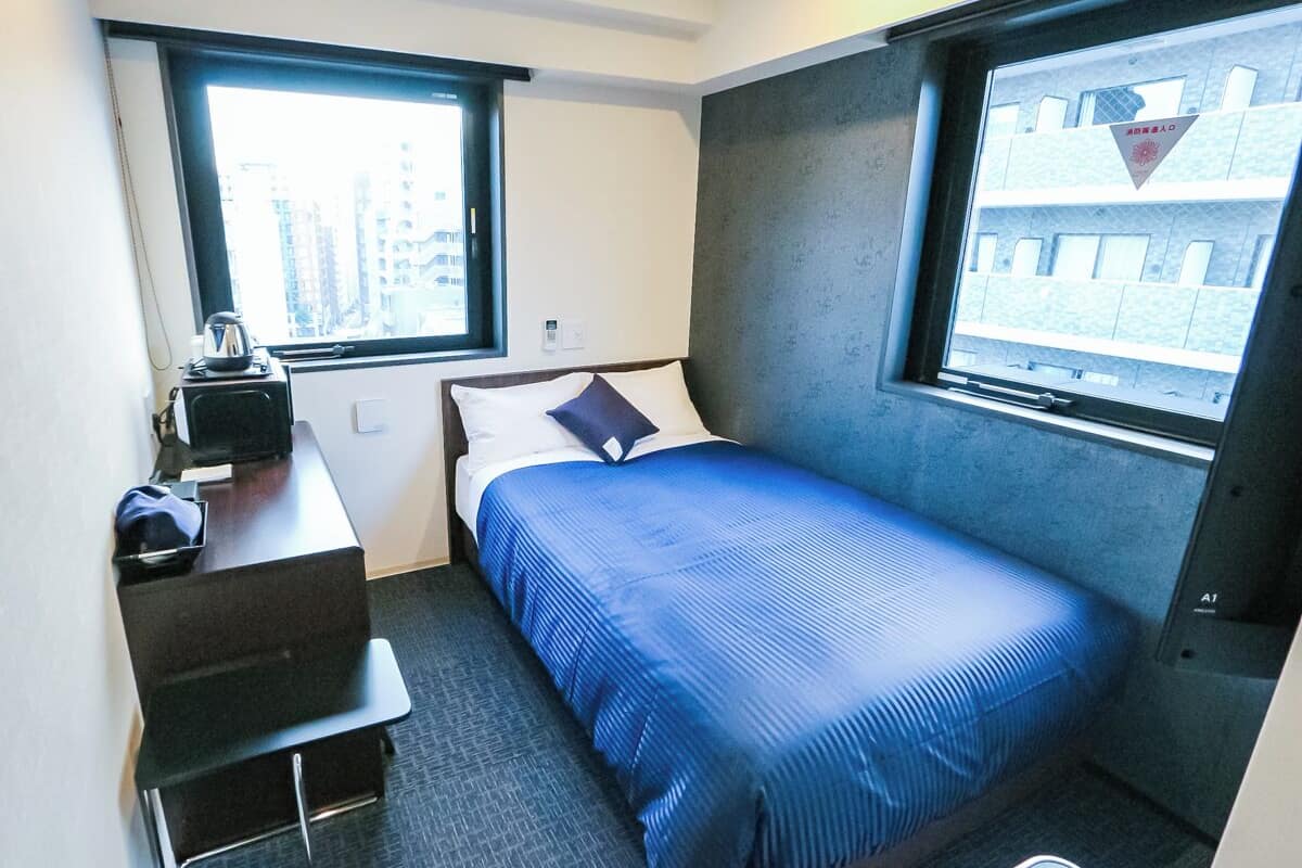 A compact hotel room with a single bed, a desk, and a chair. There are two windows providing a view of the cityscape.