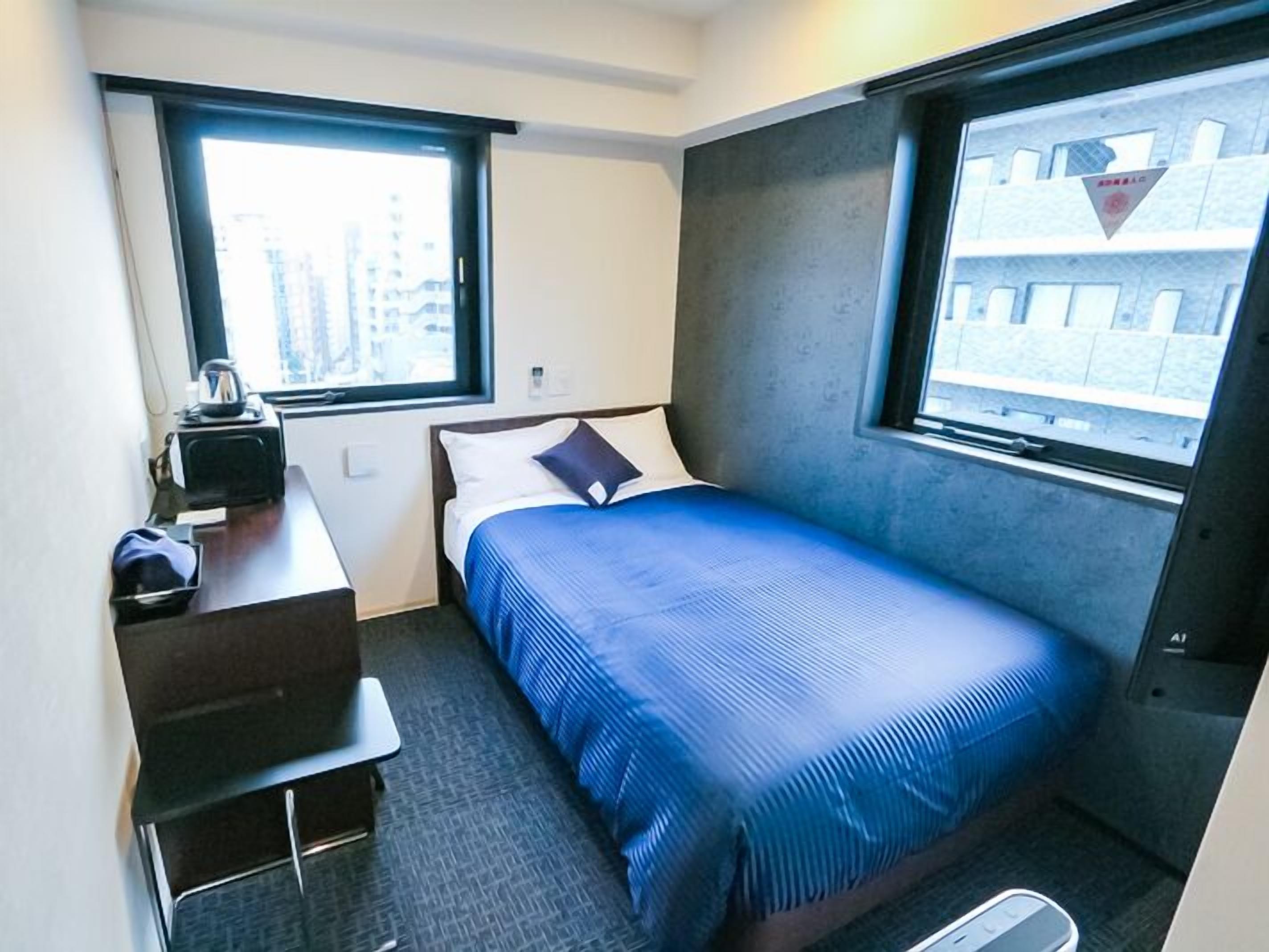 A small hotel room with a bed, a desk with a kettle, a chair, and a window.