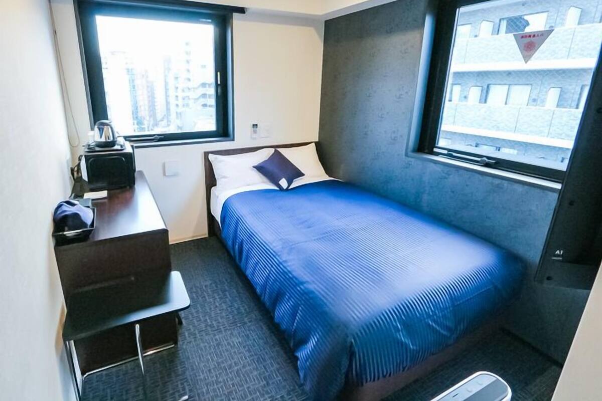 A small hotel room with a bed, a desk with a kettle, a chair, and a window.