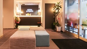 A hotel reception area with a reception desk, a large potted plant, and a seating area with two ottomans. - Hotel Livemax Tokyoshintomicho (Tokyo)