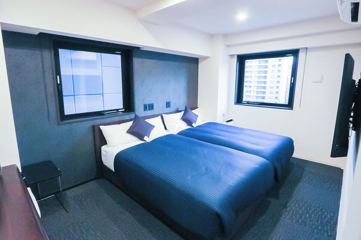 A hotel room with two beds, a small table, a TV, and windows with a view of buildings.