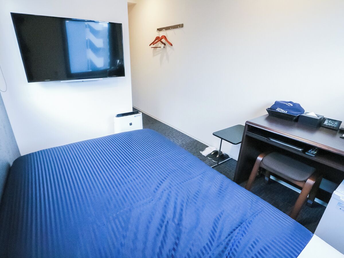 Habitación de hotel con cama, escritorio, silla, televisor montado en la pared y perchero con ganchos.