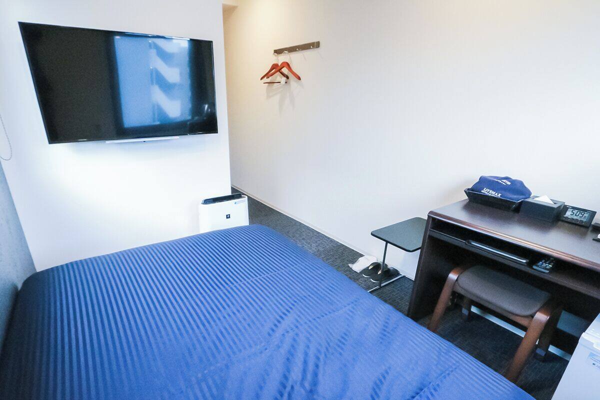 A hotel room with a bed, a desk, a chair, a TV mounted on the wall, and a coat rack with hangers.