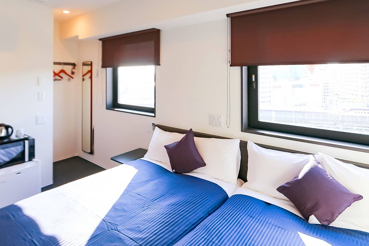 Ein Hotelzimmer mit einem Bett, zwei Fenstern mit Jalousien, einem Nachttisch und einem Spiegel.