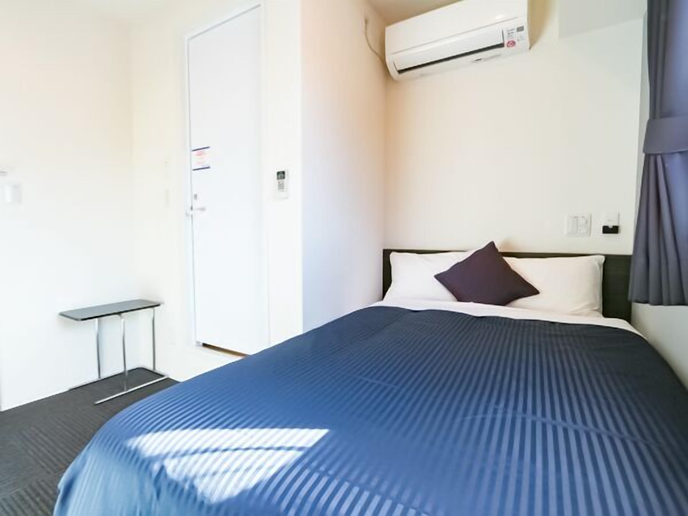 A hotel room with a bed, a small table, an air conditioning unit, and a door.