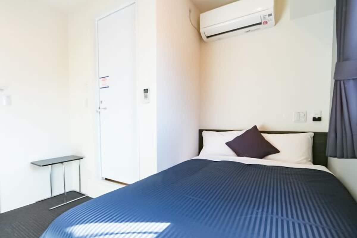 Hotellihuone, jossa on sänky, pieni pöytä, ilmastointilaite ja ovi.