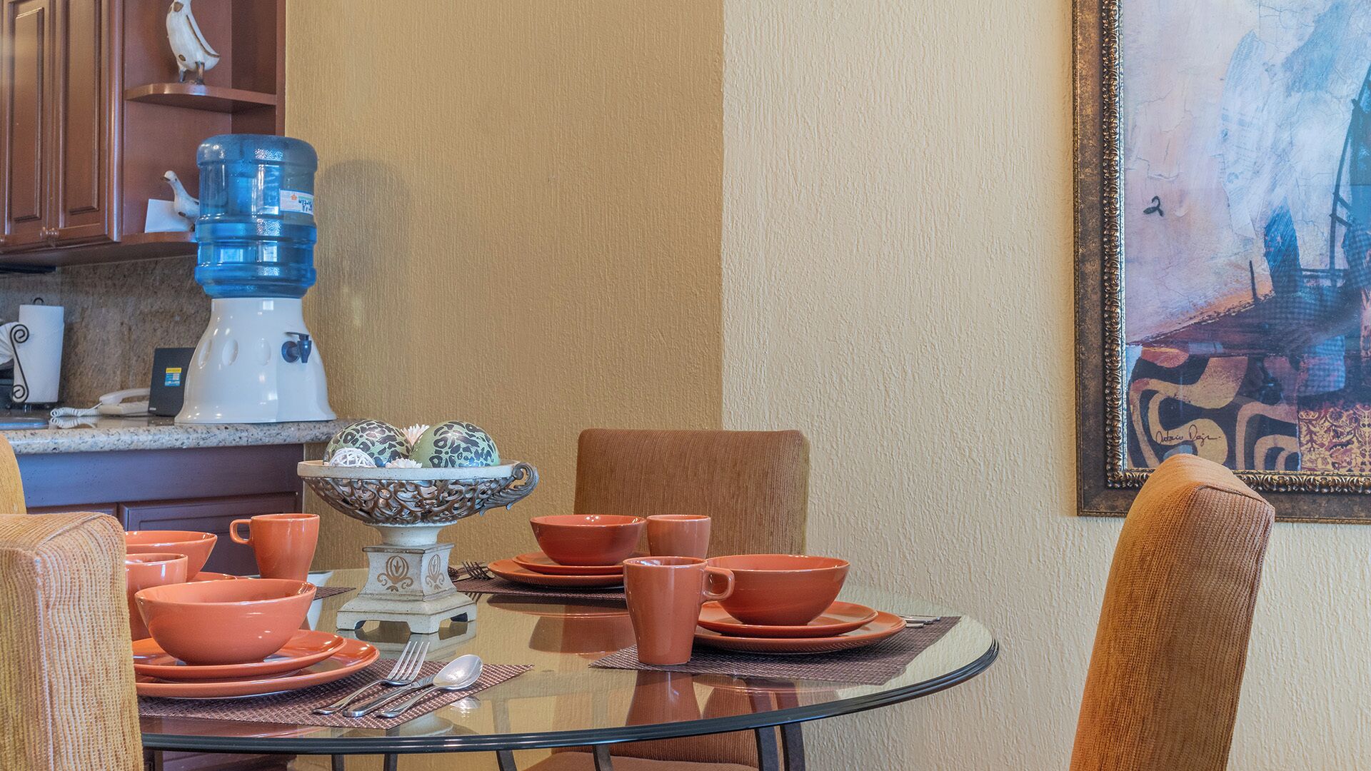 Un coin repas avec une table en verre, de la vaisselle orange et un bol décoratif.