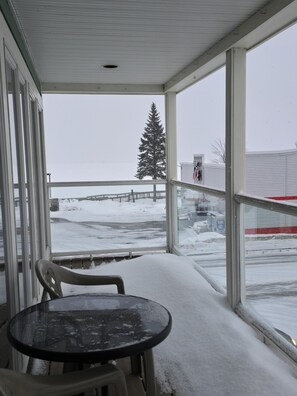 A covered outdoor seating area with a table and chairs, overlooking a snowy landscape and a distant tree.