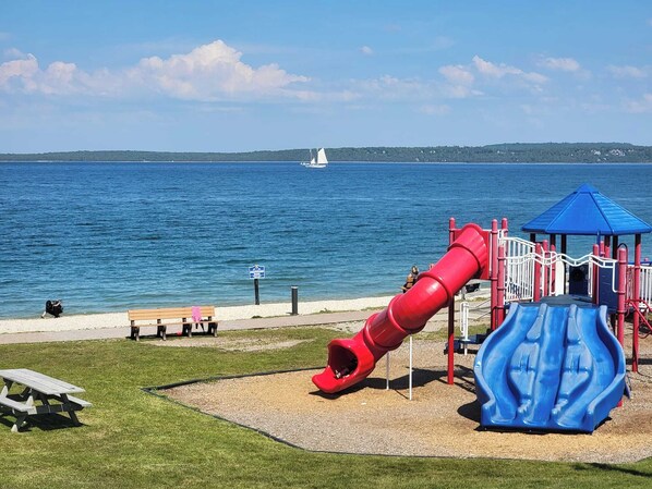 A playground with a red slide and blue slide, located near a body of water with a sailboat in the distance.