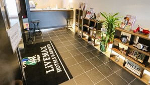 A reception area with a black and white checkered floor, a reception desk, a seating area with two chairs, a bookshelf with various items, and a sign that reads "LIVEMAX GROUP HOTELS & RESORTS".