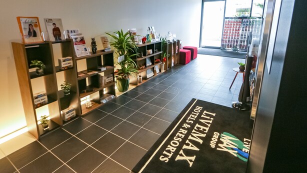 A hotel reception area with a black tiled floor, a reception desk, and a 'Live Max' sign. There are shelves with various items and a 'Hello' sign on the wall.