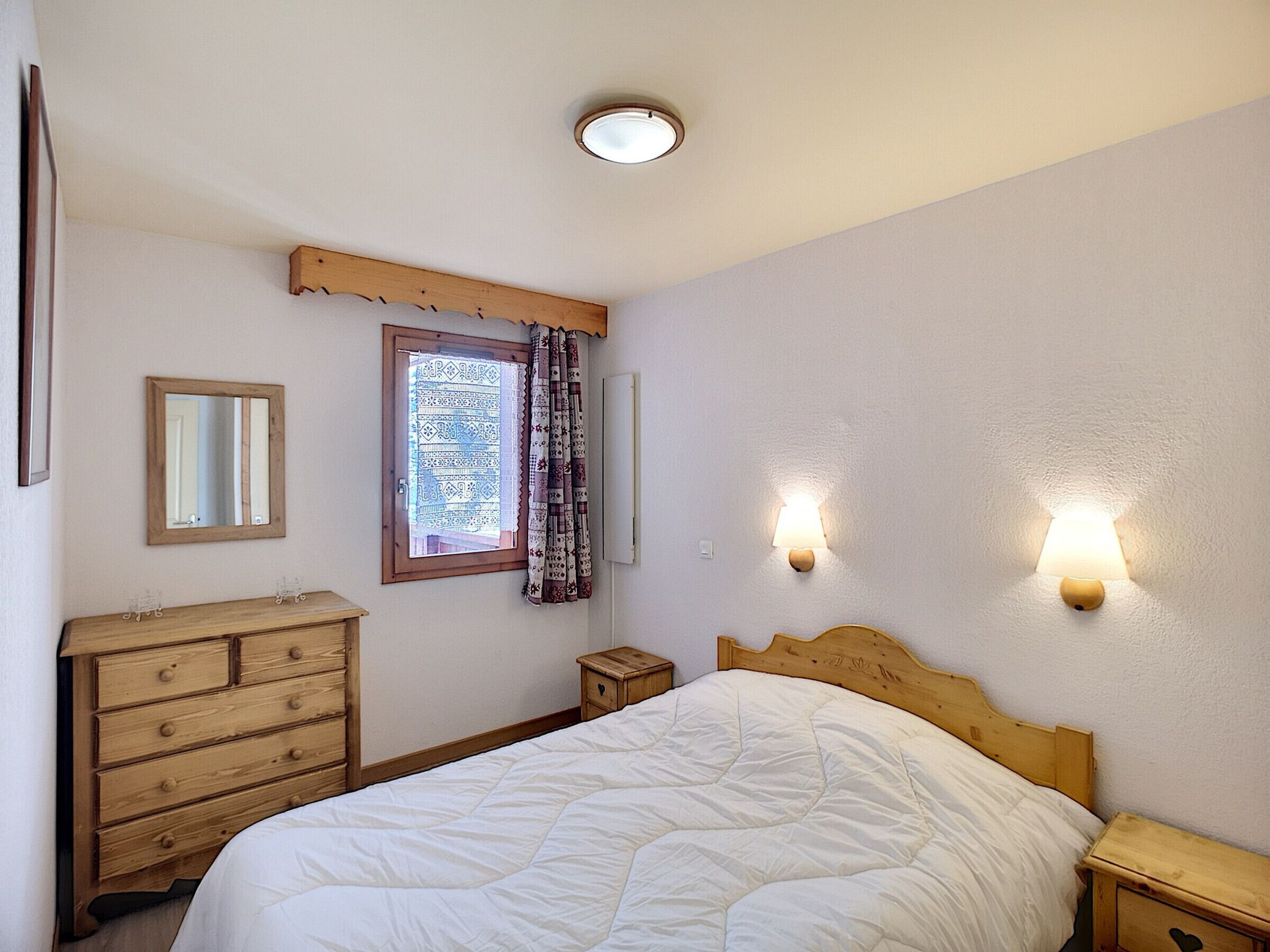 Un dormitorio con cama, cómoda de madera, mesita de noche y dos lámparas de pared.