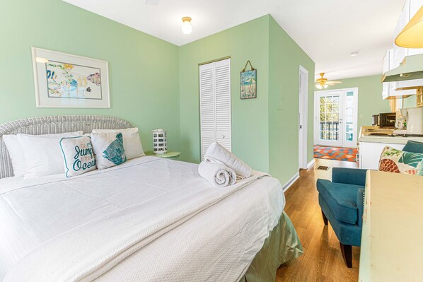 Un dormitorio con cama, almohadas, mesita de noche, pared con un cuadro enmarcado, ventana con contraventanas y puerta que da a un baño.