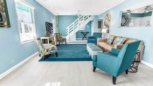 A spacious living room with a staircase, teal and white furniture, and a large window with blinds. - Coastal Tybee House & Bungalow : Steps from Beach (Tybee Island)