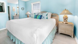 A bedroom with a large bed, a wicker nightstand, a lamp, and a window with blinds. - Coastal Tybee House & Bungalow : Steps from Beach (Tybee Island)