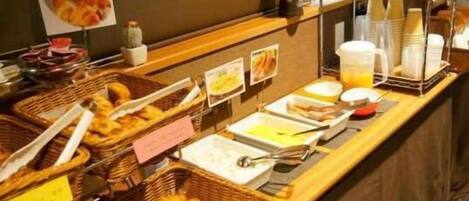 A buffet counter with various food items and utensils, including baskets of pastries and containers of beverages.