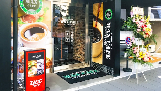 Eingang eines Cafés mit einem großen Schild mit der Aufschrift 'MAX CAFE' und einer Blumenkomposition davor.