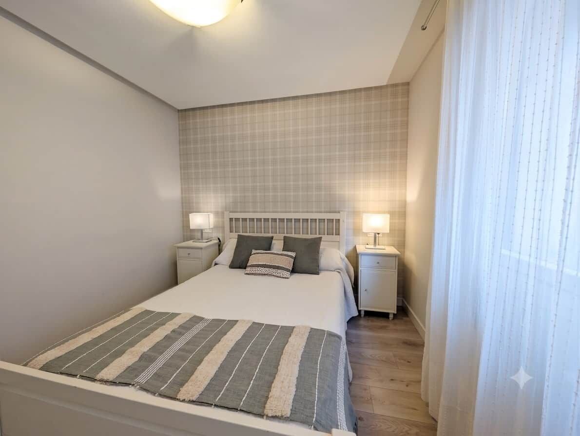 Un dormitorio con una cama, dos mesitas de noche, cabecero y una ventana con cortinas.