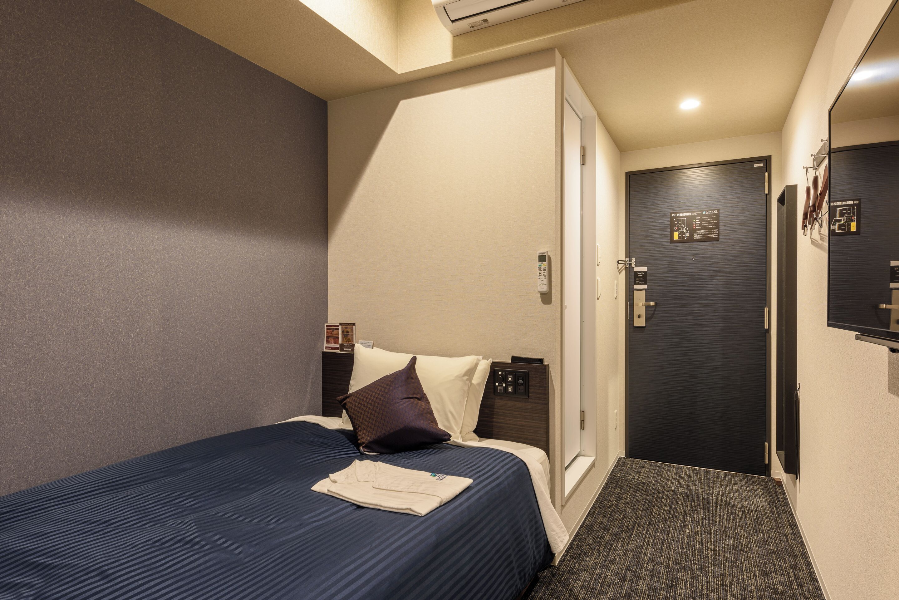 A hotel room with a bed, a bedside table, a door, and a wall-mounted air conditioning unit.