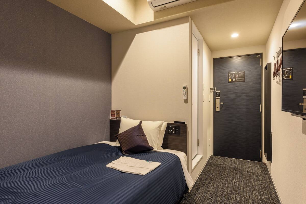 Quarto de hotel com cama, mesa de cabeceira, porta e ar condicionado fixado na parede.