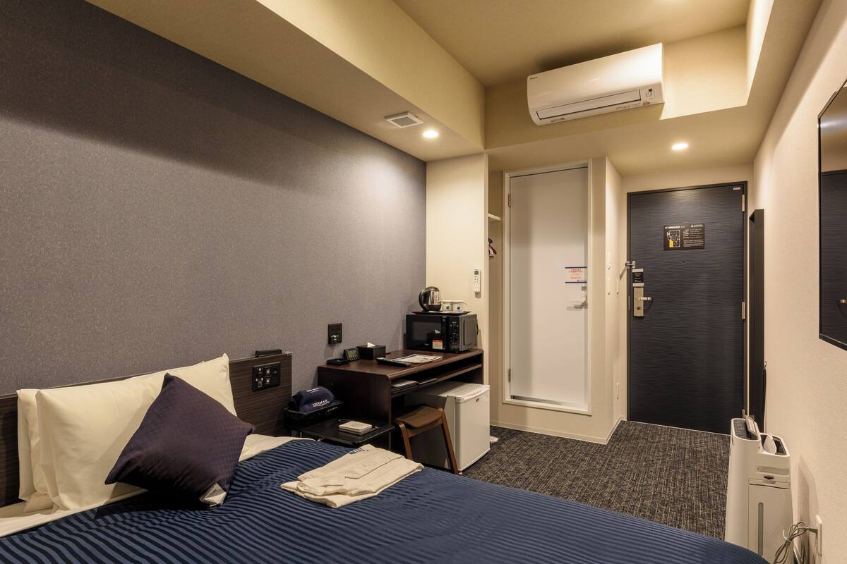 Quarto de hotel com cama, escrivaninha, cadeira, micro-ondas e pequeno frigorífico.