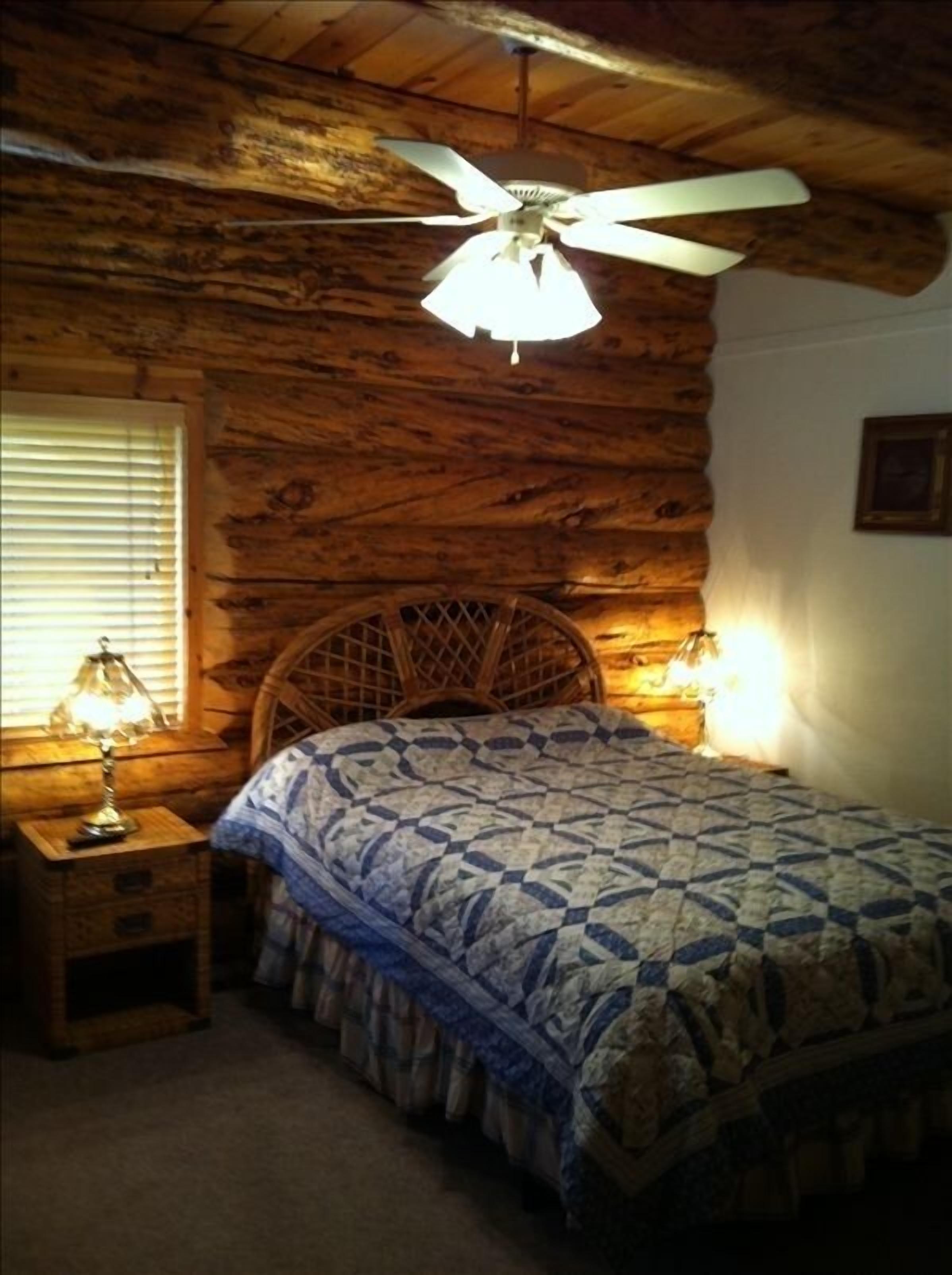 A rustic bedroom with a wooden headboard, a bed with a patterned quilt, a nightstand with a lamp, and a ceiling fan.