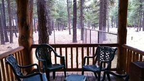 A covered outdoor seating area with wooden railings and chairs, overlooking a forested area.