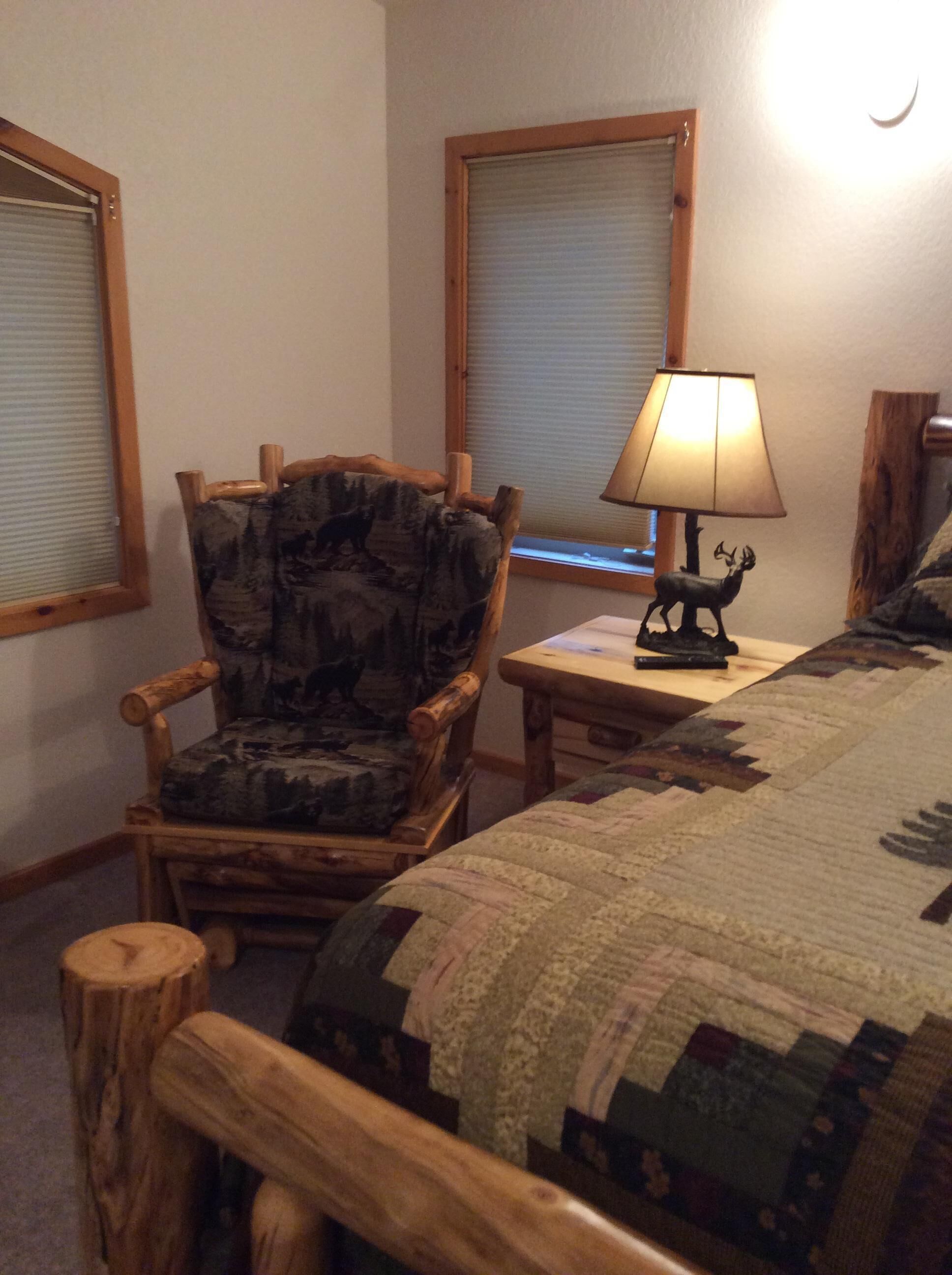 Une chambre à coucher avec un cadre de lit en bois, une courtepointe, une chaise à motifs camouflage, une lampe et deux fenêtres équipées de stores.